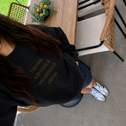 A person wearing a black crewneck sweatshirt with gold text that reads 'THRIVING SURVIVING & OCCASIONALLY CRYING' paired with blue shorts and white sneakers, photographed from an overhead angle in an outdoor patio setting.