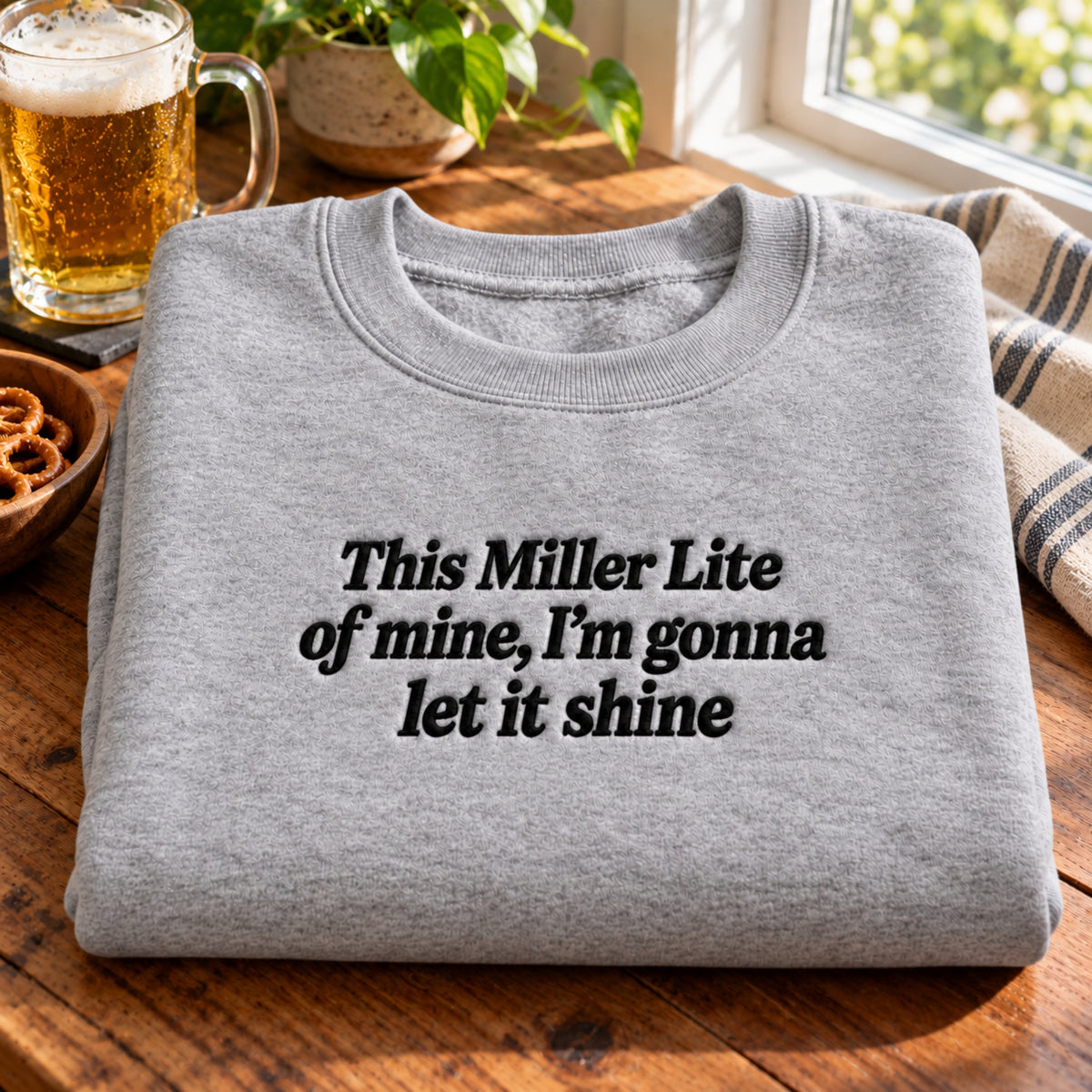 Funny beer lover sweatshirt with black embroidered text reading “This Miller Lite of mine, I’m gonna let it shine,” folded on a rustic wooden table beside a beer mug and pretzels.