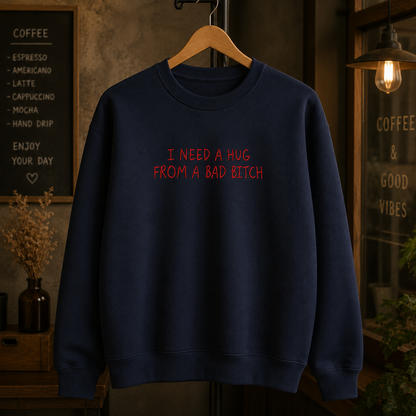 A navy blue crewneck sweatshirt hanging on a wooden hanger, featuring the text 'I NEED A HUG FROM A BAD BITCH' printed in red across the chest. The background depicts a cozy, dimly lit cafe interior with a chalkboard menu and warm hanging light.