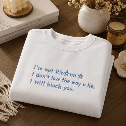 White crewneck sweatshirt neatly folded on a wooden table, embroidered with blue text reading "I'm not Rihanna I don't love the way u lie, I will block you." Surrounding the sweatshirt are a white bubble candle on books, a lit jar candle, dried flowers, and a tasseled coaster.