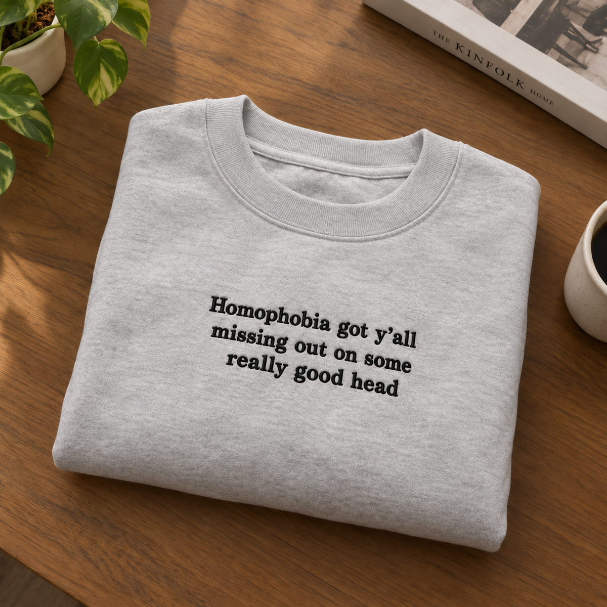 A light gray crewneck sweatshirt with black text reading 'Homophobia got y'all missing out on some really good head' is folded on a wooden table with a plant, coffee cup, and book visible in the background.