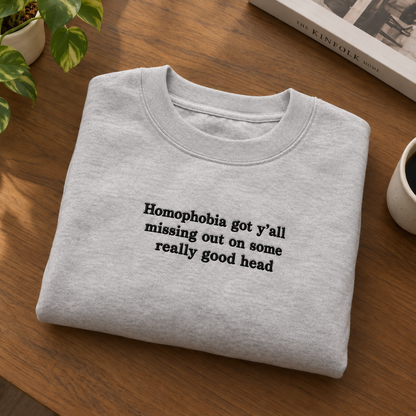 A light gray crewneck sweatshirt with black text reading 'Homophobia got y'all missing out on some really good head' is folded on a wooden table with a plant, coffee cup, and book visible in the background.