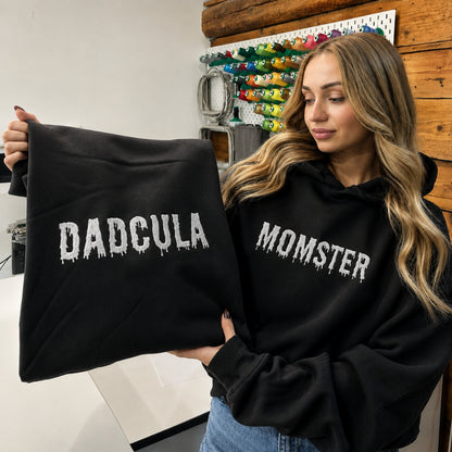 A woman wearing a black hoodie with 'MOMSTER' in white dripping text holds up a matching black hoodie with 'DADCULA' in the same white dripping font. The setting is a workshop or studio with a pegboard of colorful spools in the background.