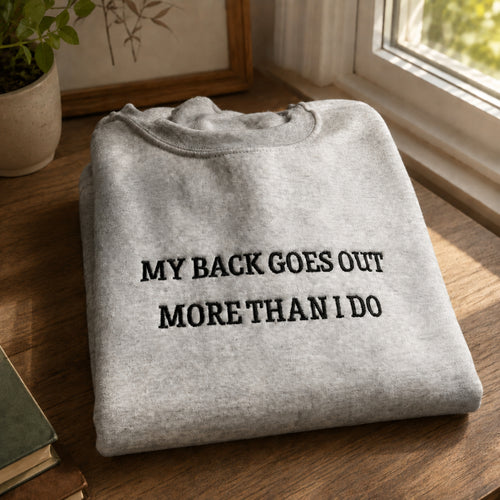 A folded light gray sweatshirt with black embroidered text reading 'MY BACK GOES OUT MORE THAN I DO' sits on a wooden table near a window, with a potted plant and framed art in the background.