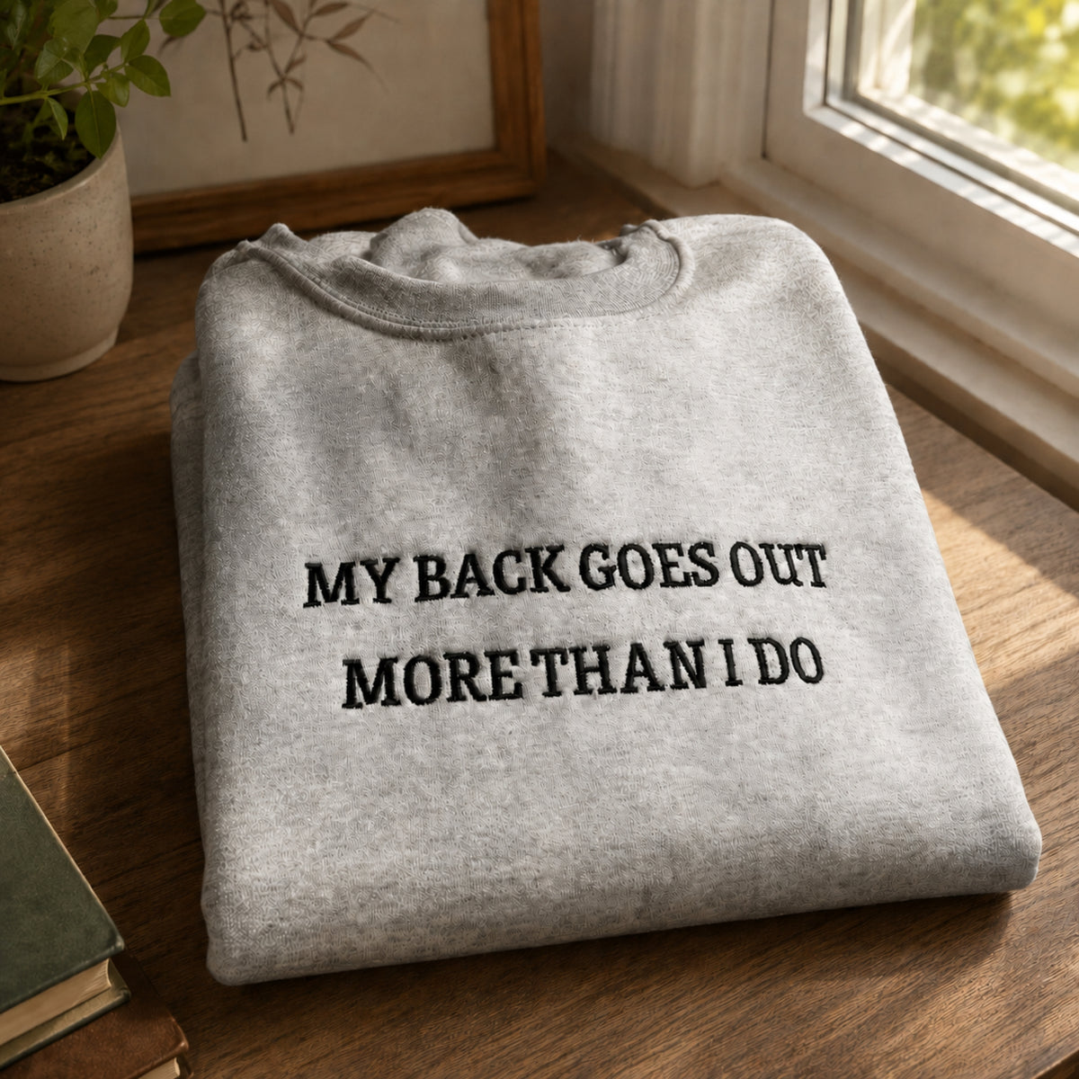 A folded light gray sweatshirt with black embroidered text reading 'MY BACK GOES OUT MORE THAN I DO' sits on a wooden table near a window, with a potted plant and framed art in the background.