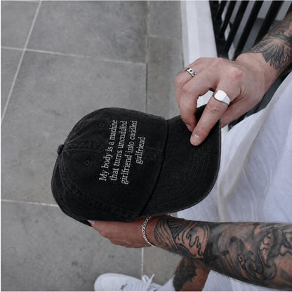 A black baseball cap with white embroidered text reading 'My body is a machine that turns unwanted girlfriend into cuddled girlfriend' being held by a tattooed person in a white shirt, standing on a gray tiled floor.