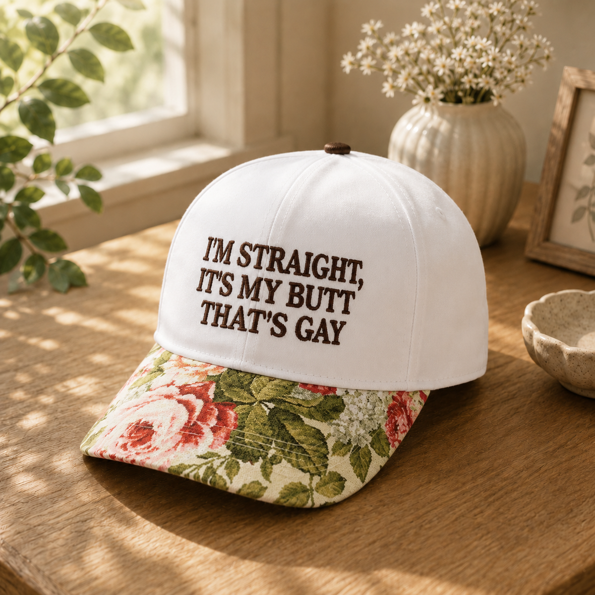 A white baseball cap with dark brown embroidered text reading 'I'M STRAIGHT, IT'S MY BUTT THAT'S GAY' sits on a wooden table. The brim has a vintage floral print with red roses and green leaves on a cream background.