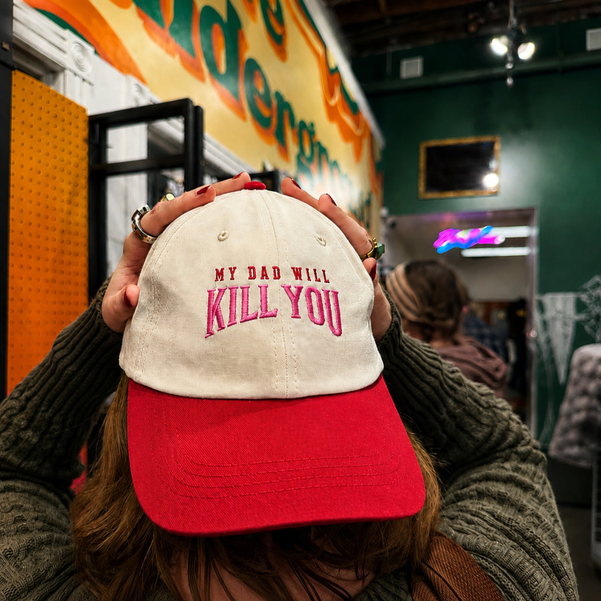 A person wearing a red and cream baseball cap with the text 'MY DAD WILL KILL YOU' in pink embroidery, holding the cap with both hands in a store setting.