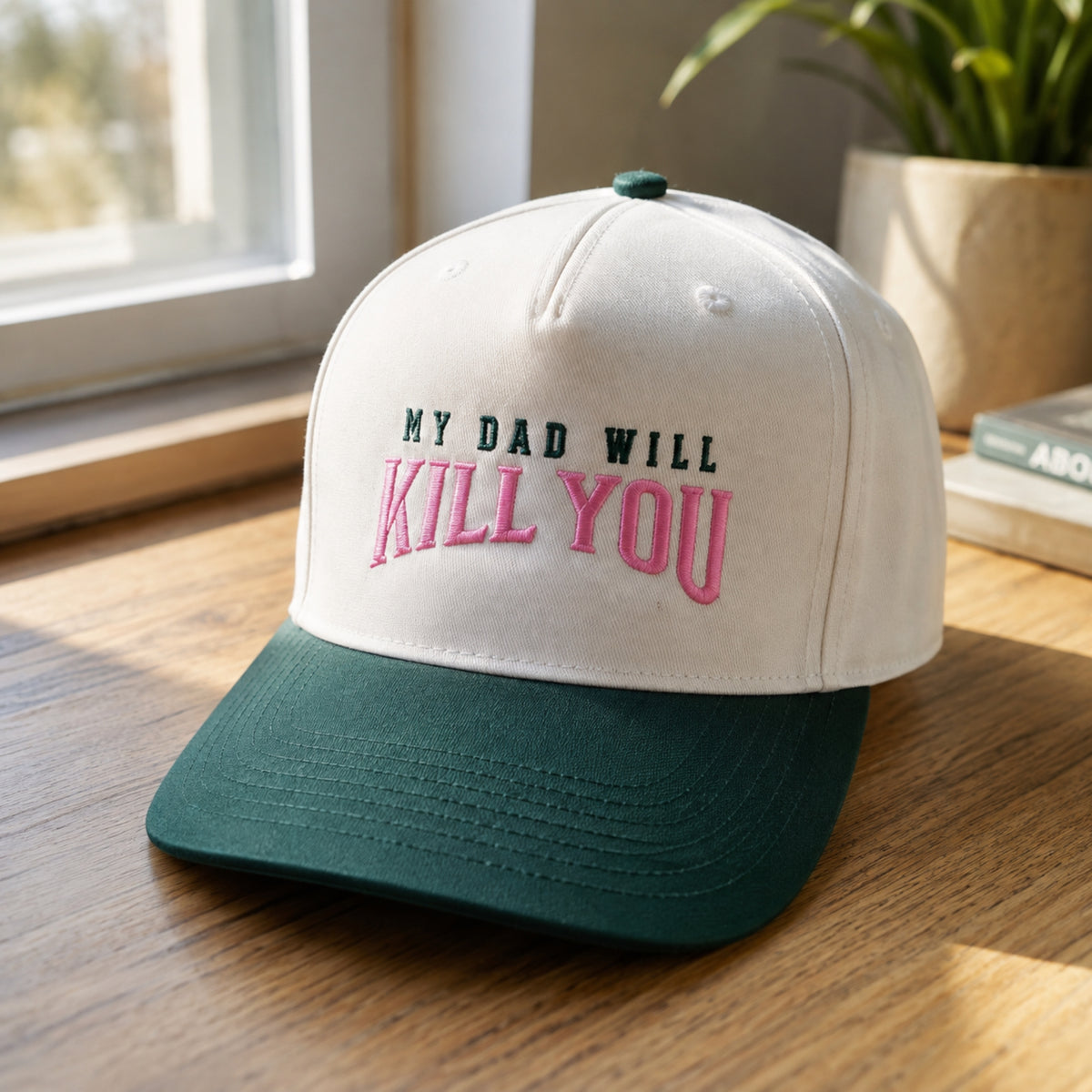 A baseball cap with a white crown and a dark green brim, featuring the embroidered text 'MY DAD WILL KILL YOU' in black and pink. The cap is styled on a wooden table near a window with a potted plant in the background.