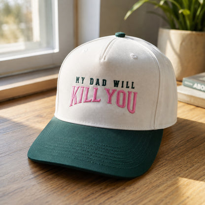 A baseball cap with a white crown and a dark green brim, featuring the embroidered text 'MY DAD WILL KILL YOU' in black and pink. The cap is styled on a wooden table near a window with a potted plant in the background.