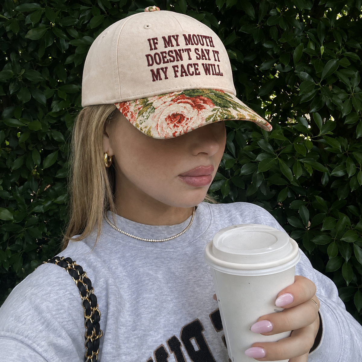 A woman wearing a beige baseball cap with dark red text reading 'IF MY MOUTH DOESN'T SAY IT MY FACE WILL' and a floral brim, holding a white coffee cup in front of a green hedge background.