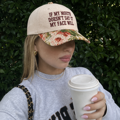 A woman wearing a beige baseball cap with dark red text reading 'IF MY MOUTH DOESN'T SAY IT MY FACE WILL' and a floral brim, holding a white coffee cup in front of a green hedge background.
