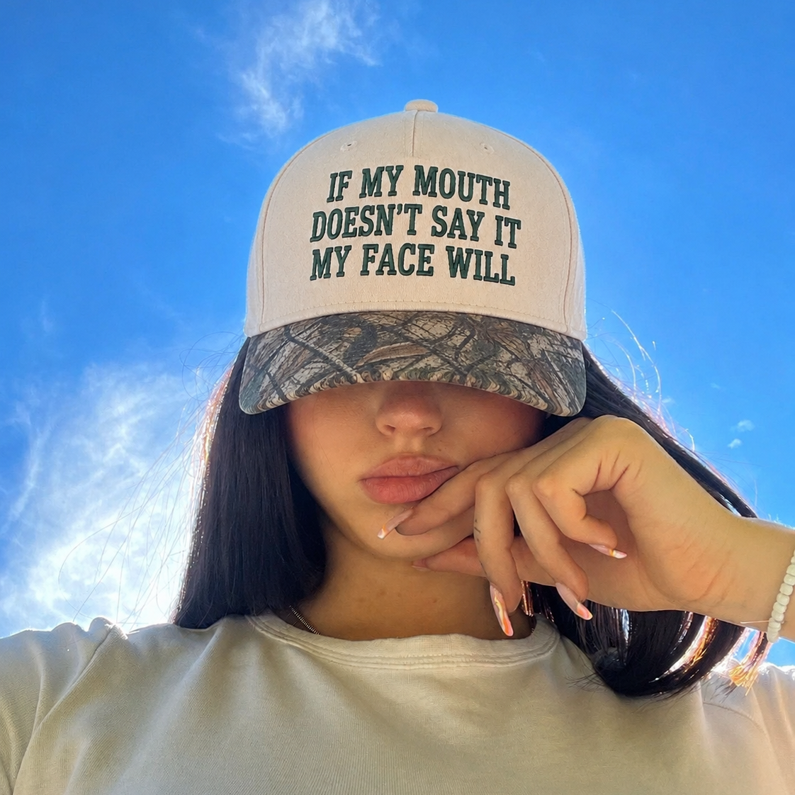 A person outdoors wearing a white trucker-style cap with dark green embroidered text reading 'IF MY MOUTH DOESN'T SAY IT MY FACE WILL' and a camouflage-patterned brim, photographed against a bright blue sky.