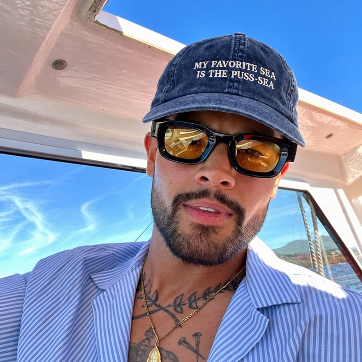 A man on a boat wearing a navy blue baseball cap with white embroidered text that reads 'MY FAVORITE SEA IS THE PUSS-SEA', black sunglasses with gold-tinted lenses, and a light blue and white striped button-down shirt.