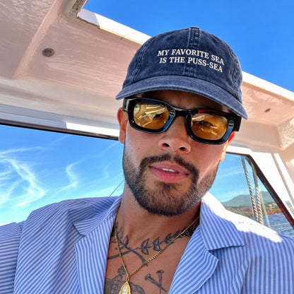 A man on a boat wearing a navy blue baseball cap with white embroidered text that reads 'MY FAVORITE SEA IS THE PUSS-SEA', black sunglasses with gold-tinted lenses, and a light blue and white striped button-down shirt.