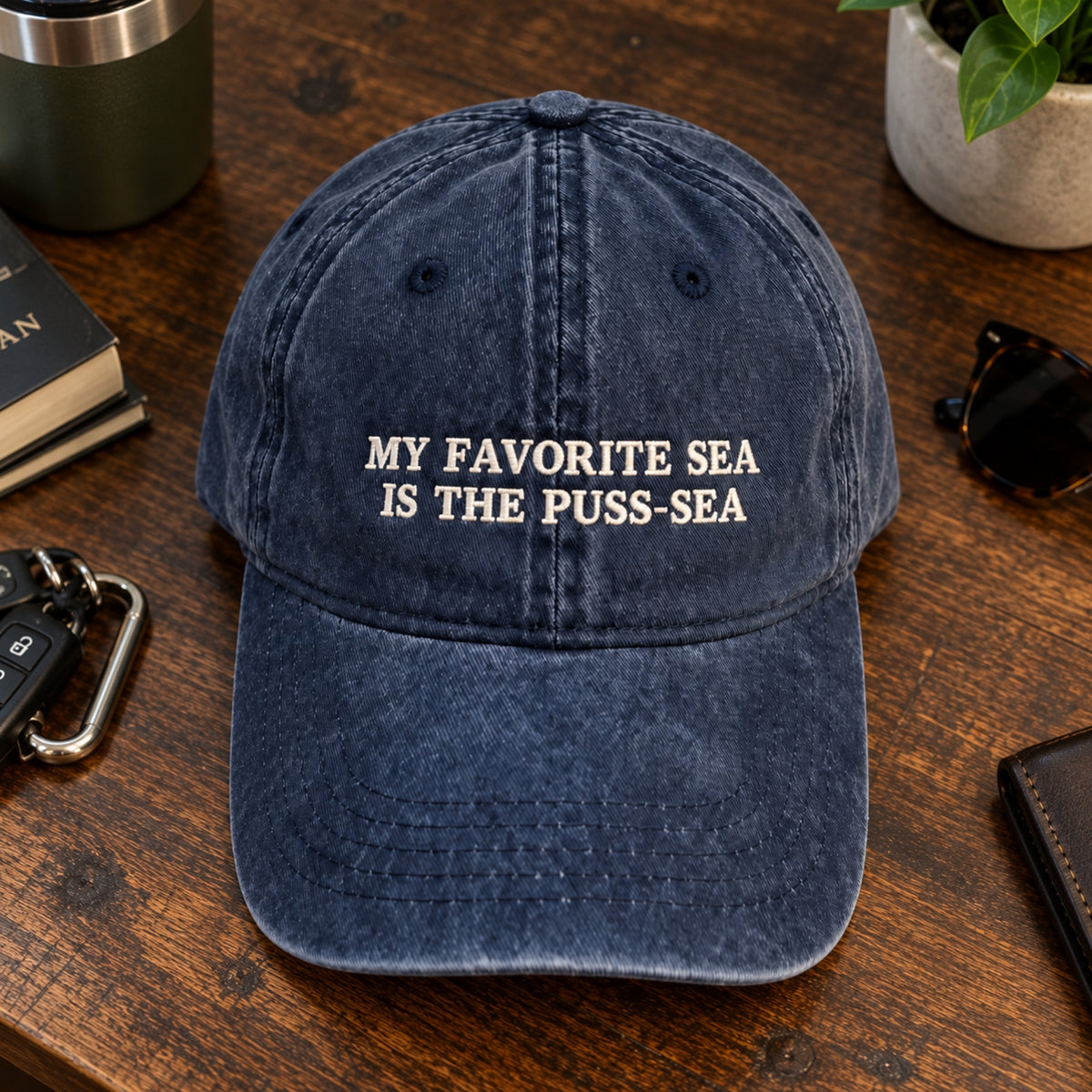 A navy blue washed baseball cap with white embroidered text reading 'MY FAVORITE SEA IS THE PUSS-SEA' sits on a wooden tabletop surrounded by everyday props like sunglasses, keys, a plant, and a book.
