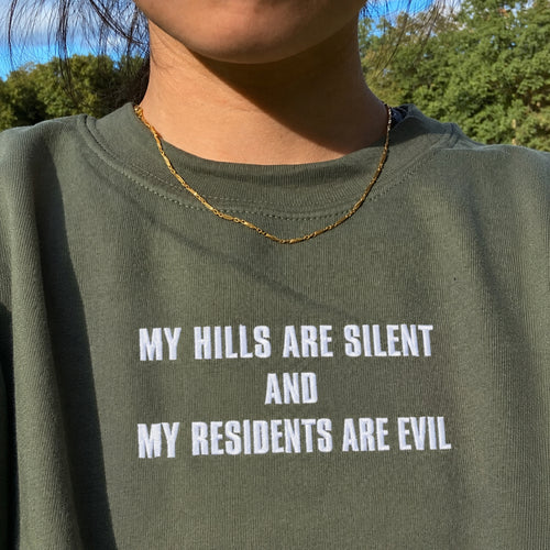 Close-up of a person wearing an olive green crewneck sweatshirt with bold white text that reads 'MY HILLS ARE SILENT AND MY RESIDENTS ARE EVIL'. The photo is taken outdoors with trees and blue sky visible in the background, and the person is also wearing a thin gold chain necklace.