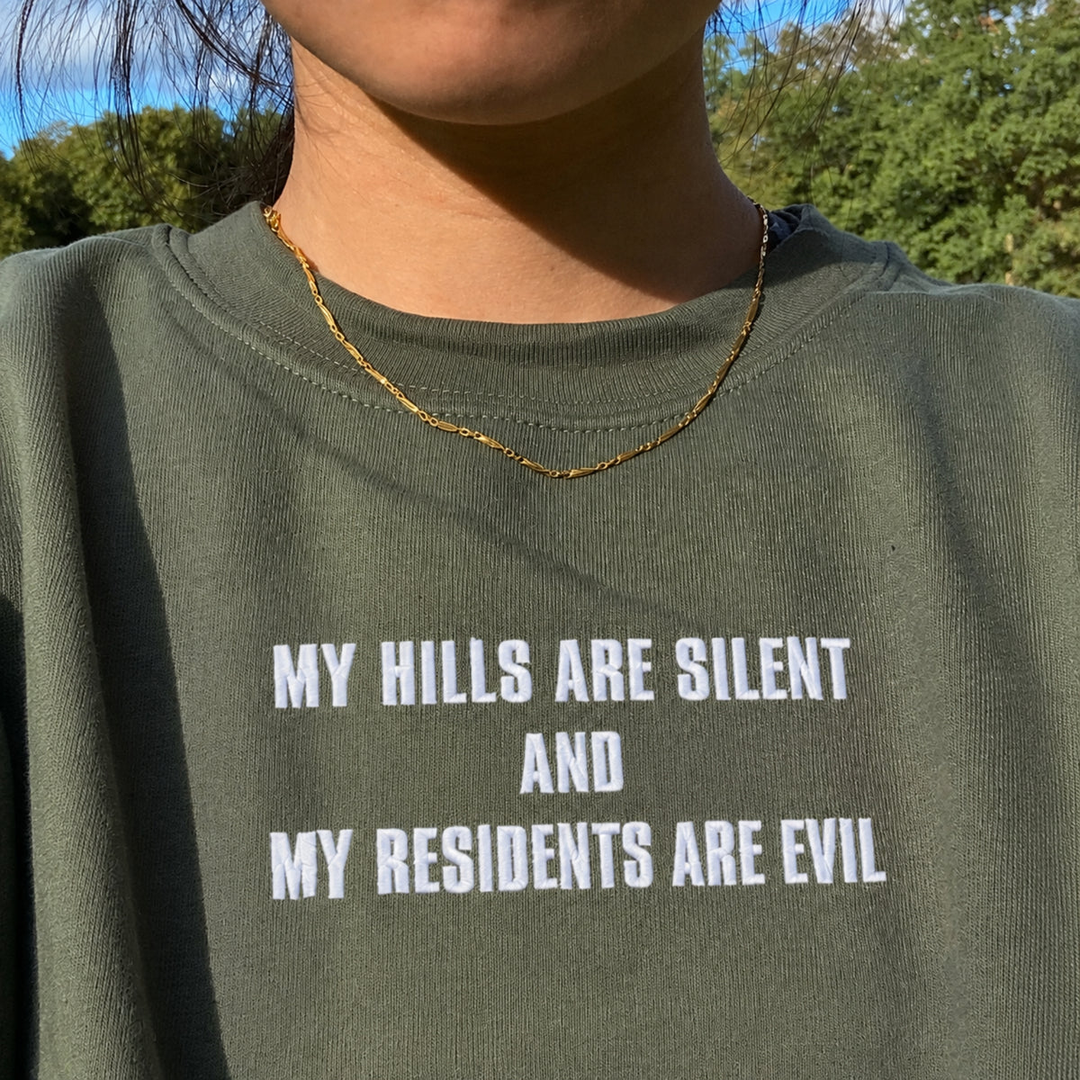 Close-up of a person wearing an olive green crewneck sweatshirt with bold white text that reads 'MY HILLS ARE SILENT AND MY RESIDENTS ARE EVIL'. The photo is taken outdoors with trees and blue sky visible in the background, and the person is also wearing a thin gold chain necklace.
