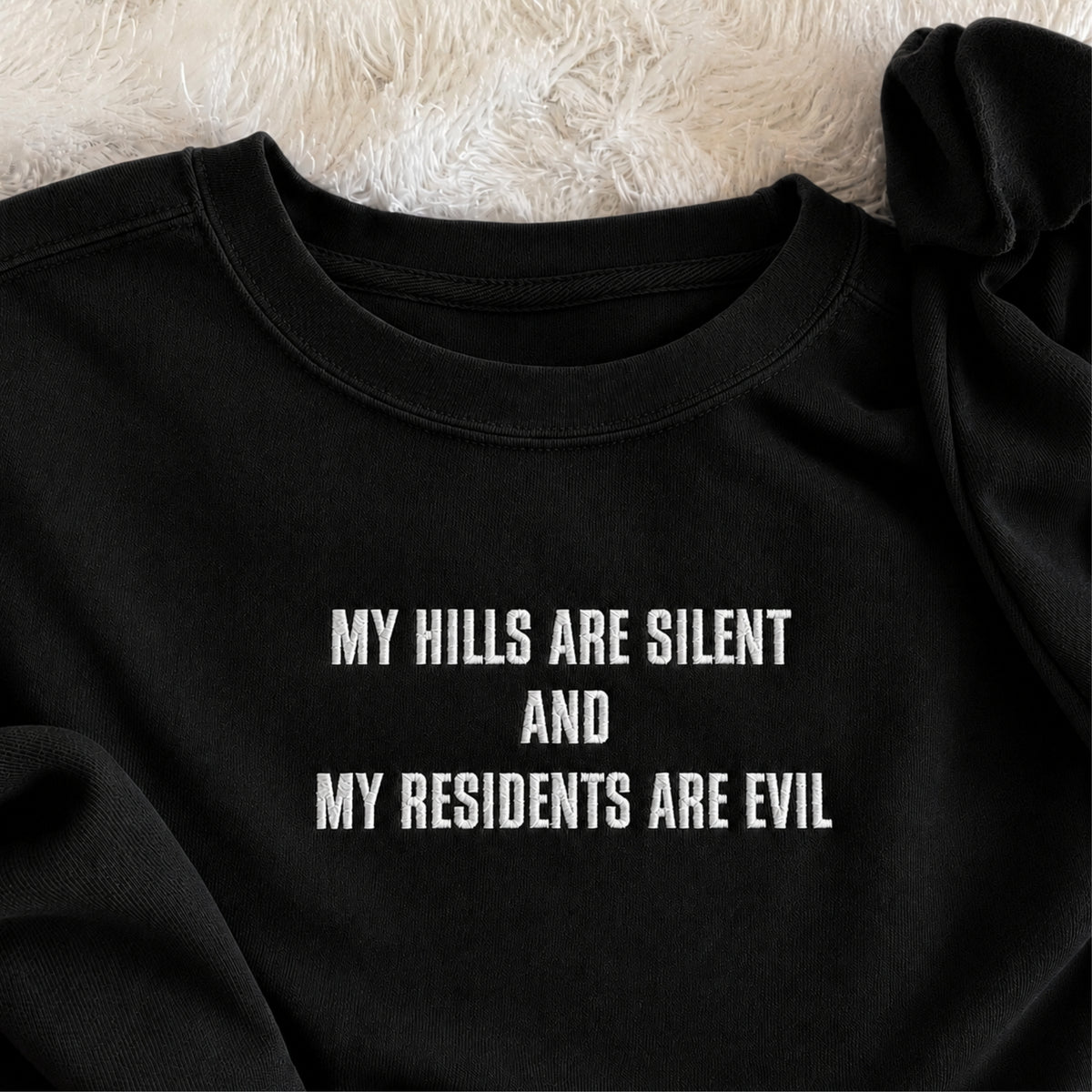 A close-up of a black crewneck sweatshirt with white embroidered text reading 'MY HILLS ARE SILENT AND MY RESIDENTS ARE EVIL', laid flat on a white faux fur surface.