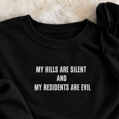 A close-up of a black crewneck sweatshirt with white embroidered text reading 'MY HILLS ARE SILENT AND MY RESIDENTS ARE EVIL', laid flat on a white faux fur surface.