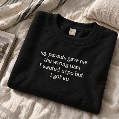 A folded black crewneck sweatshirt with white embroidered text reading 'my parents gave me the wrong tism I wanted nepo but I got au', styled on a textured cream fabric surface with a striped throw blanket and a book edge visible.