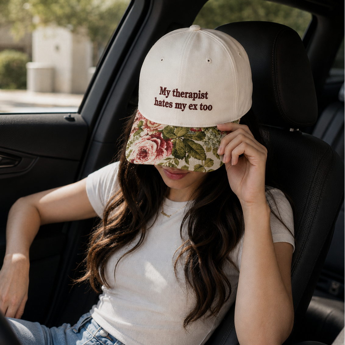Woman sitting in a car wearing a white tee and a beige embroidered baseball cap reading "My therapist hates my ex too" with a vintage floral brim.