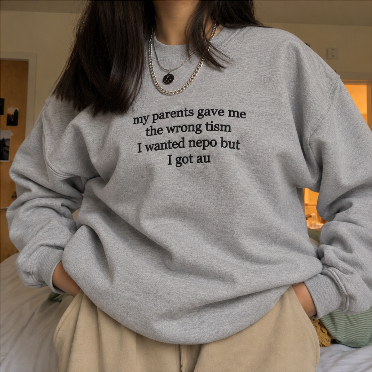 A person wearing a light gray crewneck sweatshirt with black text that reads 'my parents gave me the wrong tism I wanted nepo but I got au', paired with beige pants and layered silver necklaces.
