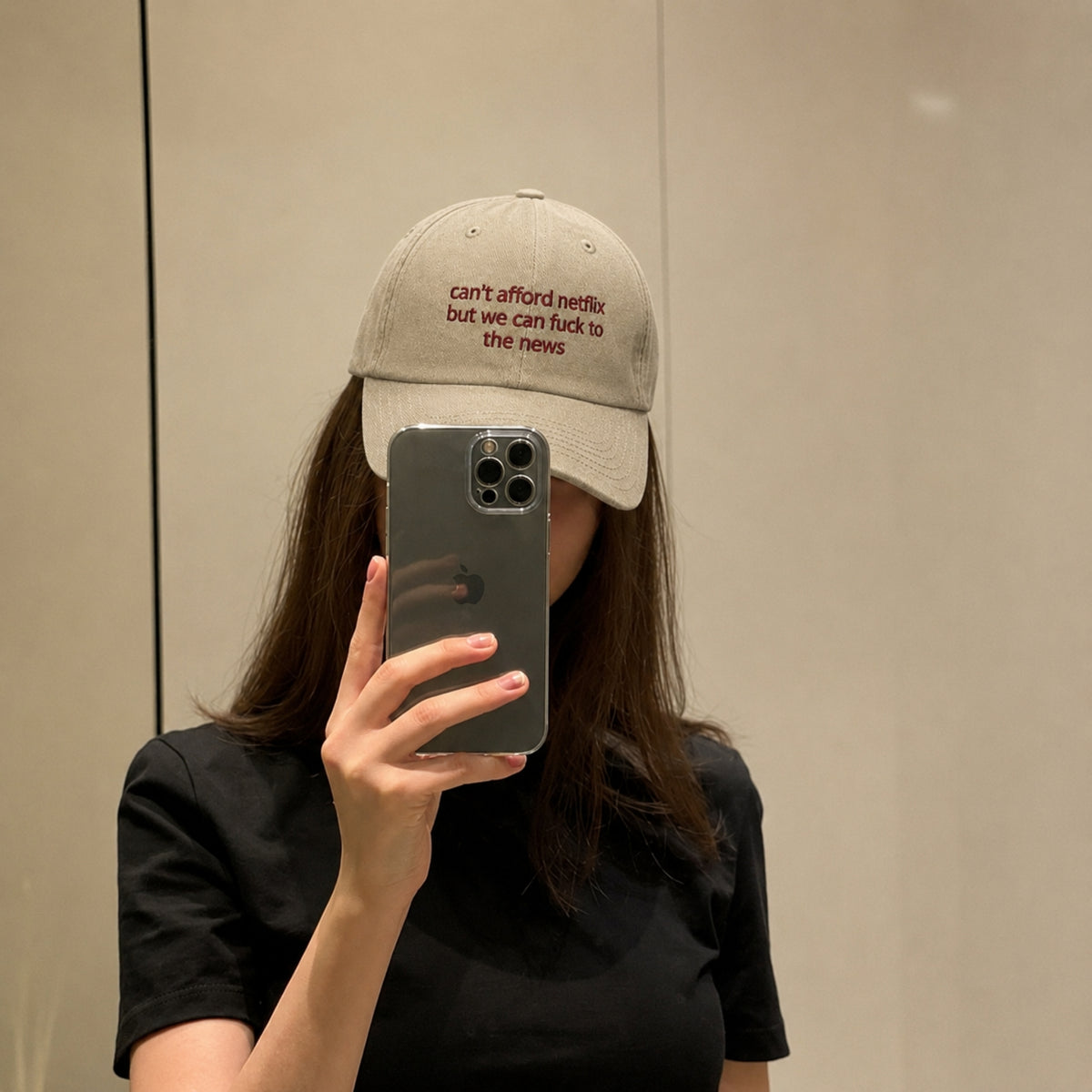A person taking a mirror selfie wearing a beige baseball cap with red embroidered text that reads 'can't afford netflix but we can fuck to the news'.