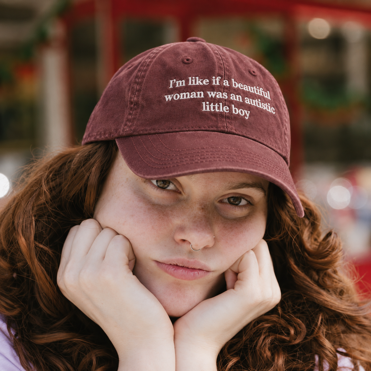 A close-up lifestyle shot of a woman with curly red hair resting her chin on her hands, wearing a maroon baseball cap with white embroidered text that reads, 'I'm like if a beautiful woman was an autistic little boy'.