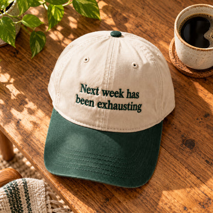 Cream and dark green embroidered baseball cap featuring “Next week has been exhausting” text, displayed on a wooden table beside coffee and a green plant.