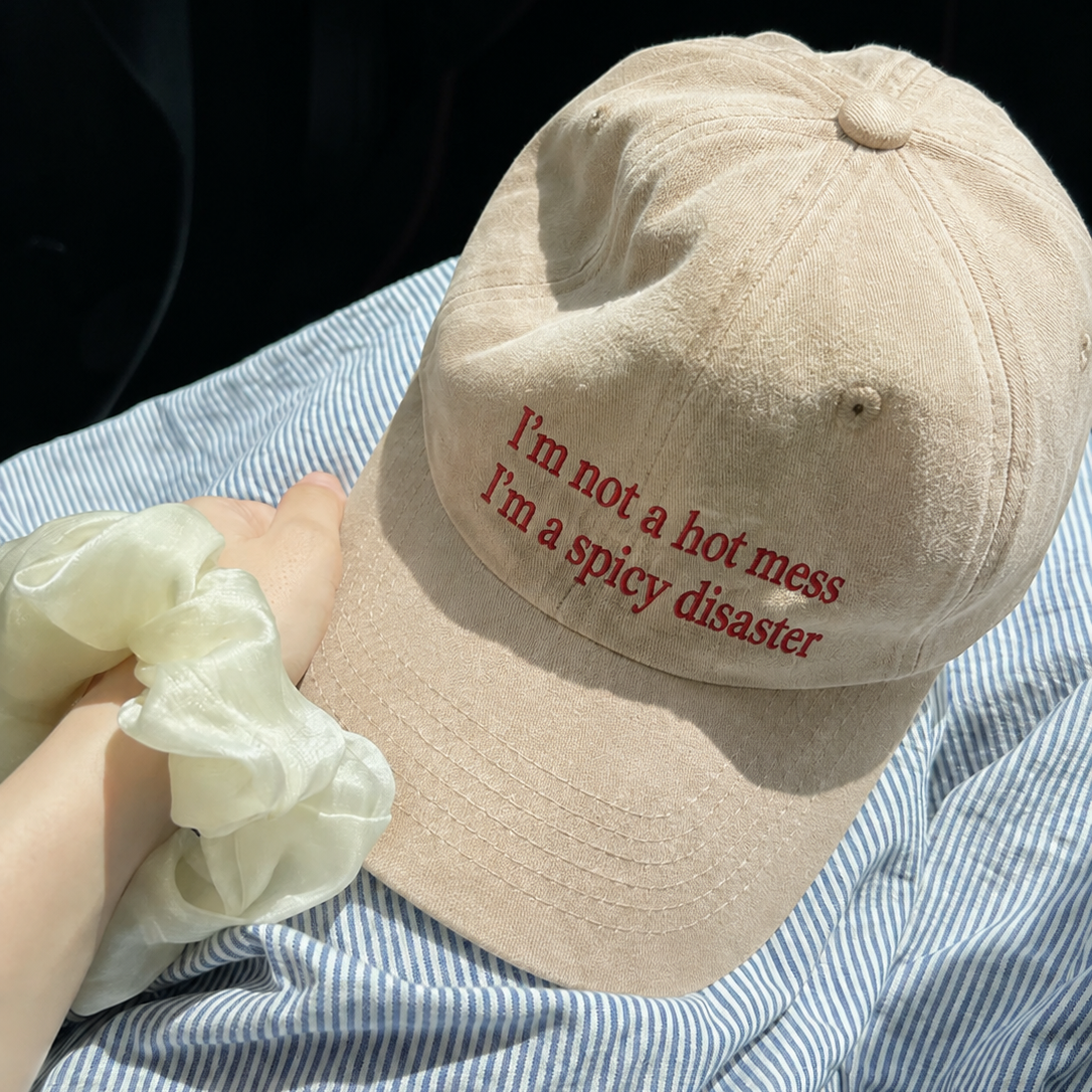 A beige baseball cap with red embroidered text reading 'I'm not a hot mess I'm a spicy disaster', resting on a blue and white striped shirt with a hand holding a white scrunchie in the foreground.