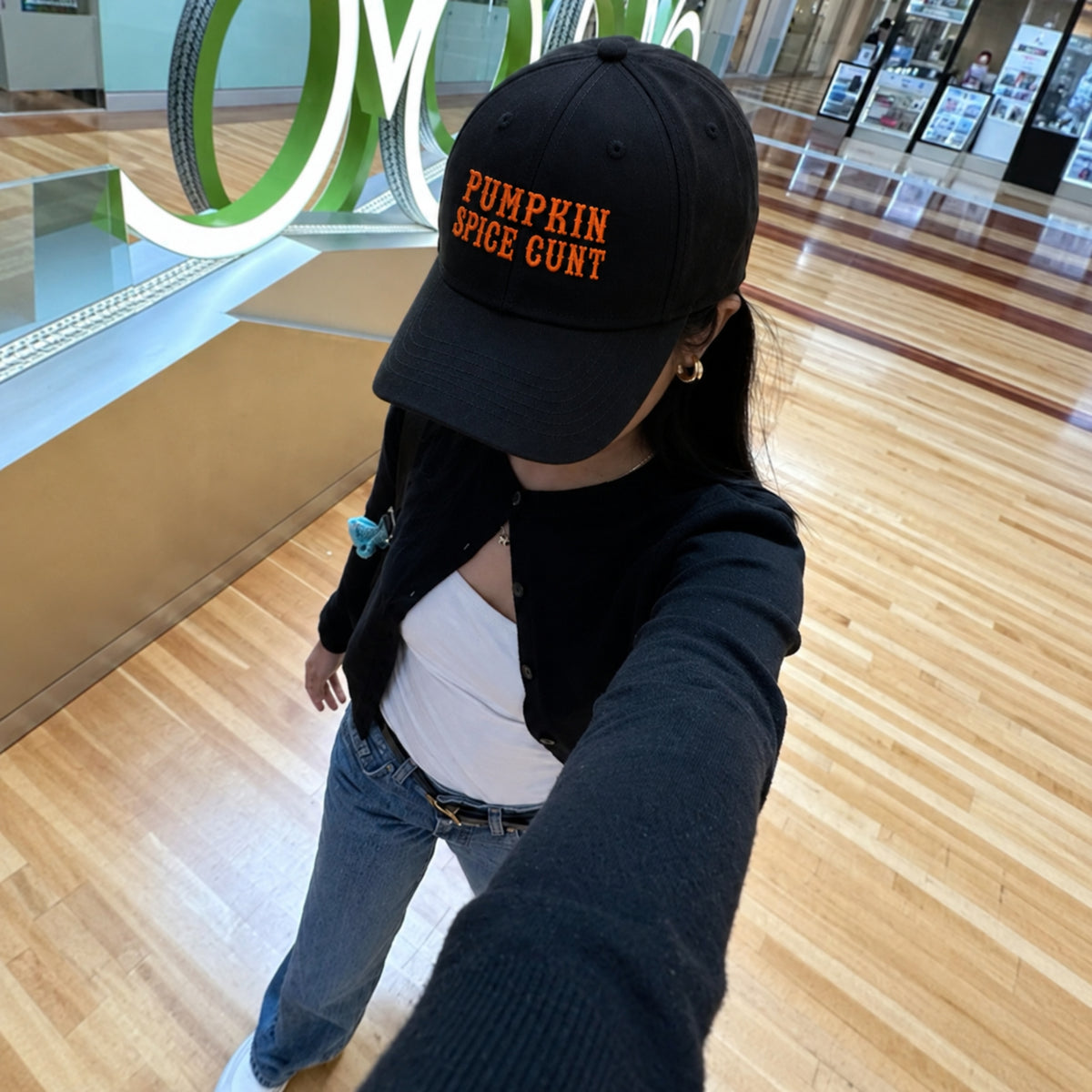 A person taking a selfie in a mall wearing a black baseball cap with orange embroidered text that reads 'PUMPKIN SPICE GUN'T'.
