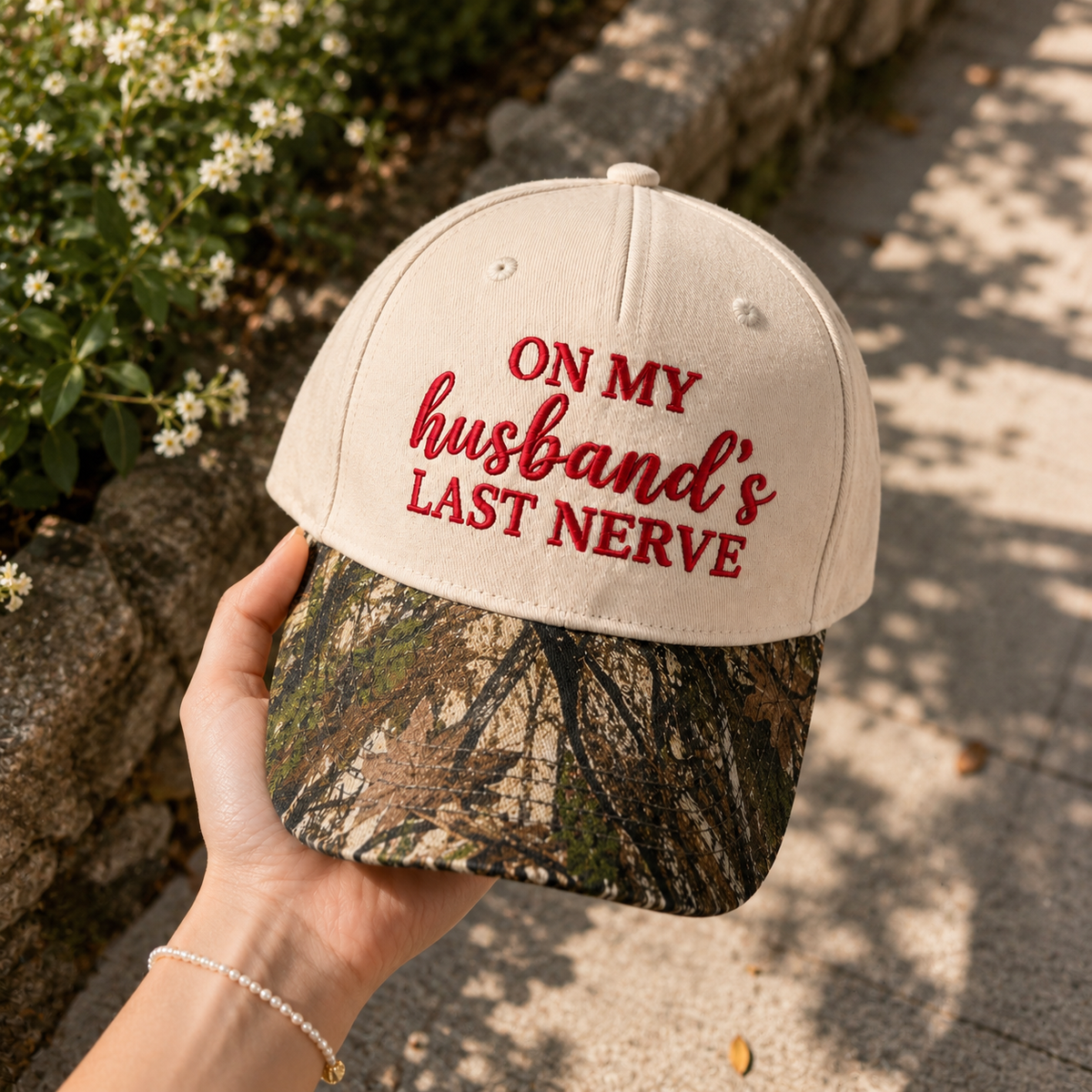 A hand holds a baseball cap outdoors. The cap has an off-white crown with red embroidered text reading 'ON MY husband's LAST NERVE' and a camouflage-patterned visor.