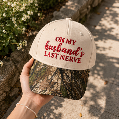 A hand holds a baseball cap outdoors. The cap has an off-white crown with red embroidered text reading 'ON MY husband's LAST NERVE' and a camouflage-patterned visor.