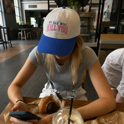 A person sitting at a cafe table wearing a white and blue baseball cap with the text 'MY DAD WILL KILL YOU' in blue and pink embroidery, looking down at a phone.