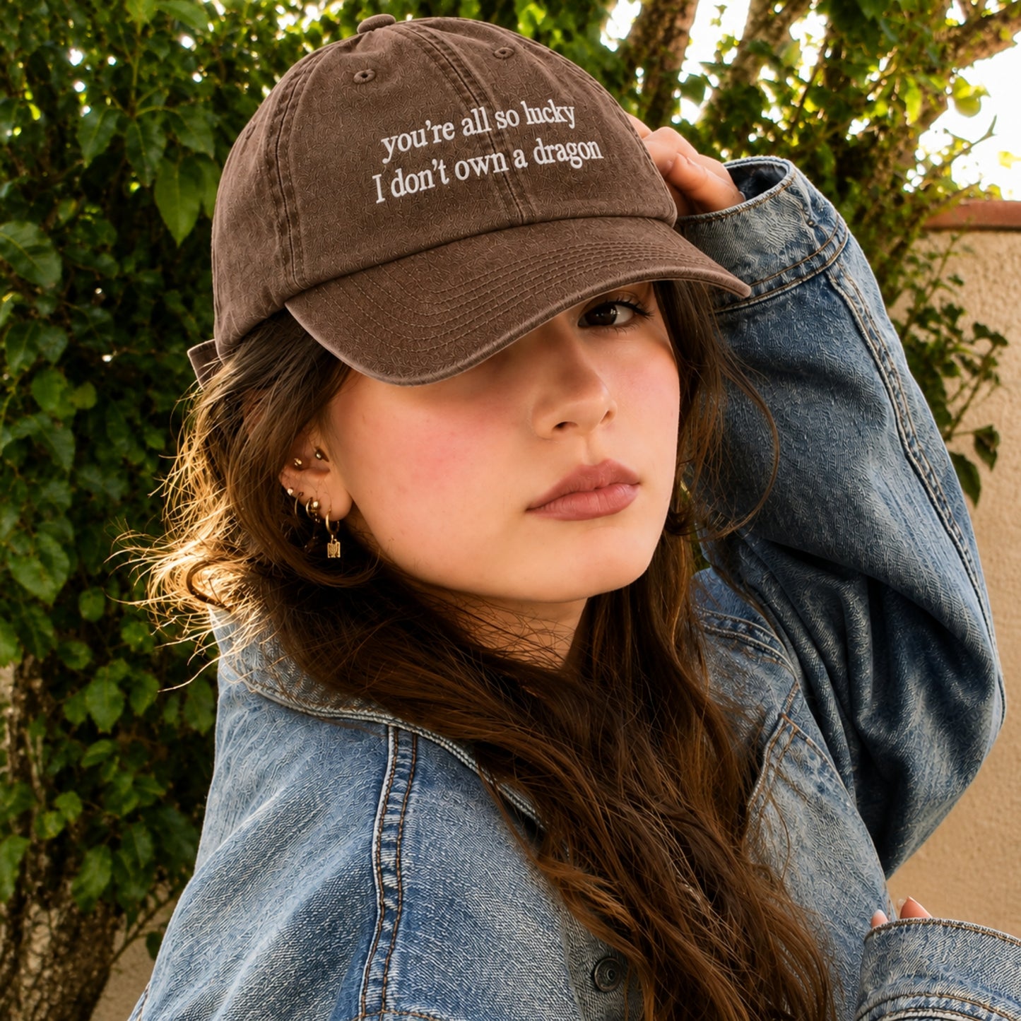 A woman wearing a brown baseball cap with white embroidered text that reads 'you're all so lucky I don't own a dragon', paired with a blue denim jacket, photographed outdoors against a background of green foliage.