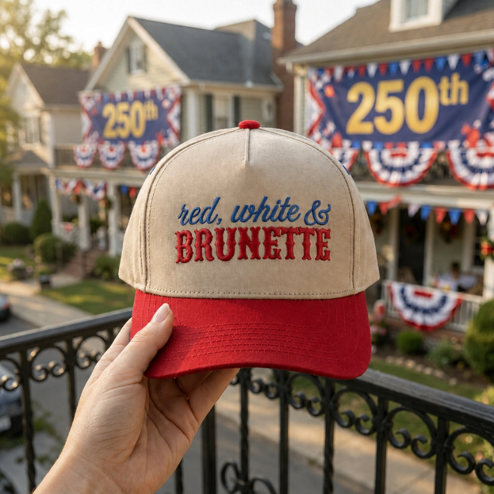 A hand holds a beige and red baseball cap in front of a decorated suburban house with patriotic banners and a large '250th' sign in the background. The cap has embroidered text reading 'red, white & BRUNETTE'.