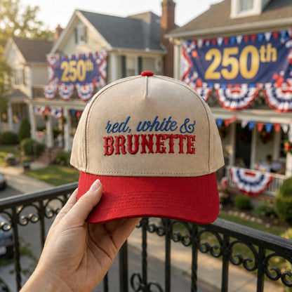 A hand holds a beige and red baseball cap in front of a decorated suburban house with patriotic banners and a large '250th' sign in the background. The cap has embroidered text reading 'red, white & BRUNETTE'.