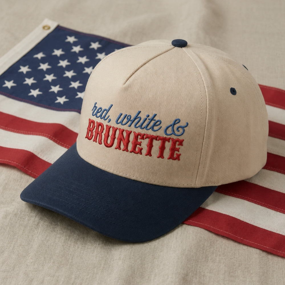 A baseball cap with a cream crown, navy blue brim, and blue and red embroidered text reading 'red, white & BRUNETTE', styled on top of an American flag against a neutral fabric background.