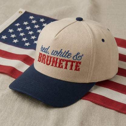 A baseball cap with a cream crown, navy blue brim, and blue and red embroidered text reading 'red, white & BRUNETTE', styled on top of an American flag against a neutral fabric background.