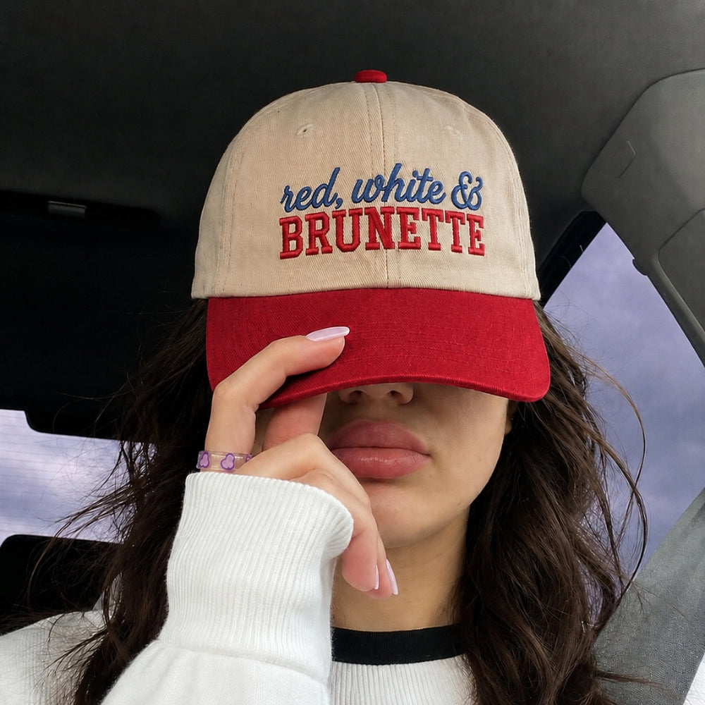 A person wearing a beige and red baseball cap with 'red, white & BRUNETTE' embroidered on the front, sitting inside a car and covering one eye with the cap brim.