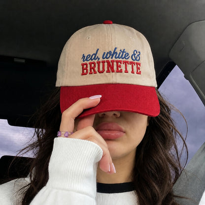 A person wearing a beige and red baseball cap with 'red, white & BRUNETTE' embroidered on the front, sitting inside a car and covering one eye with the cap brim.