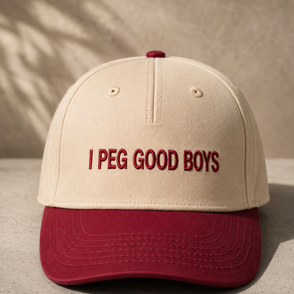 A structured baseball cap with a cream-colored crown and a dark red brim, featuring the embroidered text 'I PEG GOOD BOYS' in dark red on the front. The cap is photographed on a textured concrete surface with soft leaf shadows cast across it.