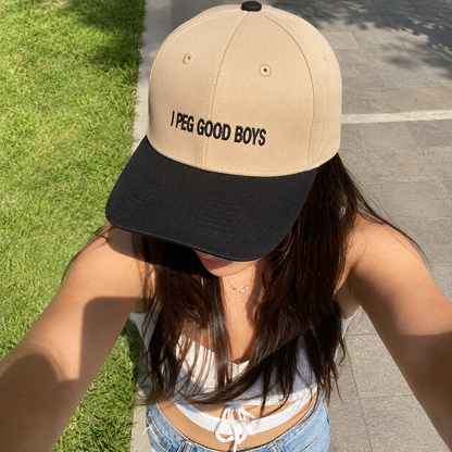 A woman wearing a beige and black baseball cap with the text 'I PEG GOOD BOYS' printed in black on the front, photographed outdoors on a paved path next to green grass.