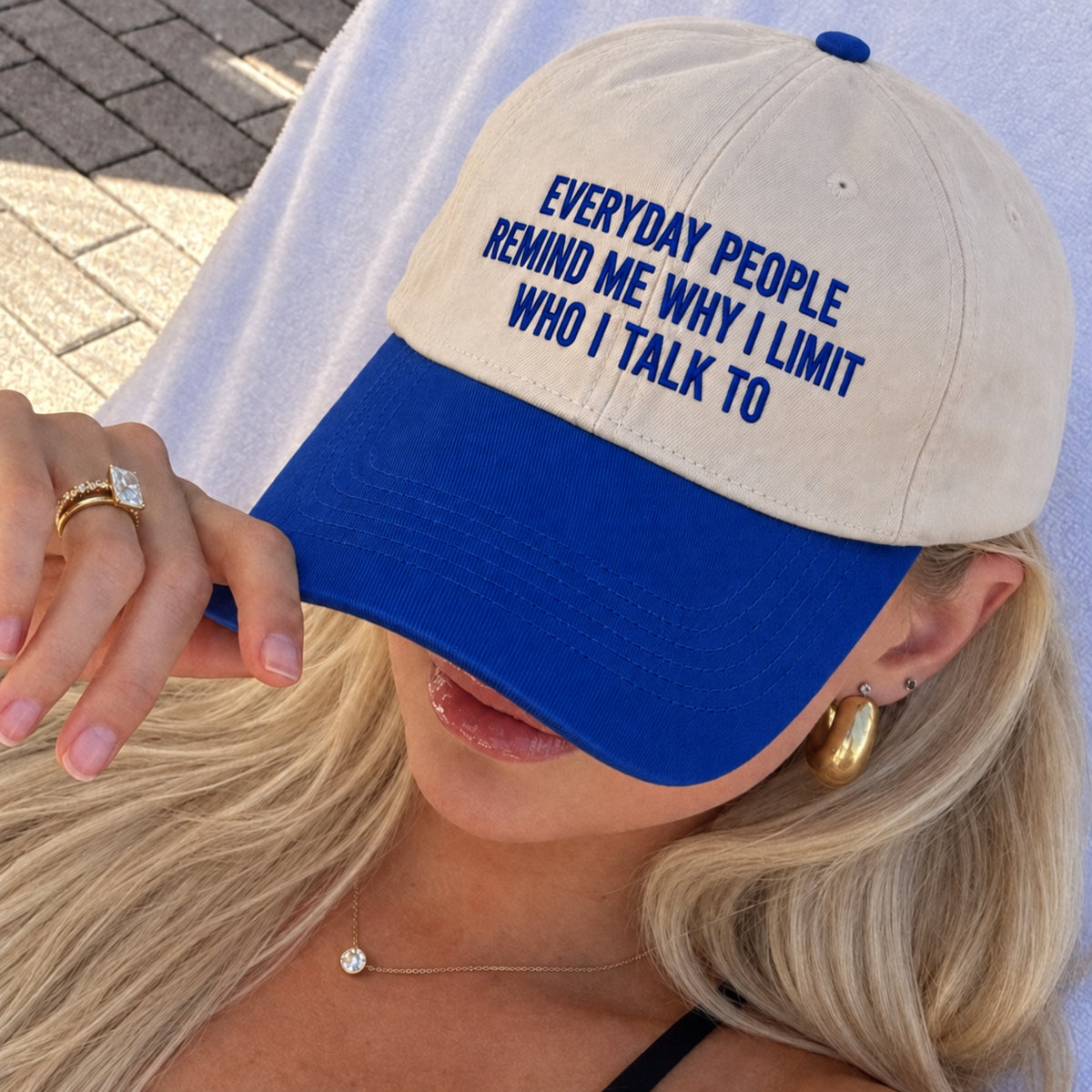 A close-up lifestyle shot of a woman wearing a beige and royal blue baseball cap. The cap features the embroidered text 'EVERYDAY PEOPLE REMIND ME WHY I LIMIT WHO I TALK TO' in blue on the front panel.