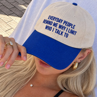 A close-up lifestyle shot of a woman wearing a beige and royal blue baseball cap. The cap features the embroidered text 'EVERYDAY PEOPLE REMIND ME WHY I LIMIT WHO I TALK TO' in blue on the front panel.