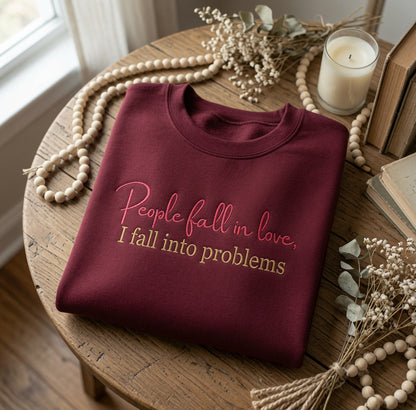 Maroon crewneck sweatshirt folded neatly on a round wooden table, embroidered with pink cursive text reading "People fall in love." above gold block lettering reading "I fall into problems." Surrounding the sweatshirt are decorative wooden beads with tassels, dried white baby's breath flowers, vintage books, and a lit jar candle.