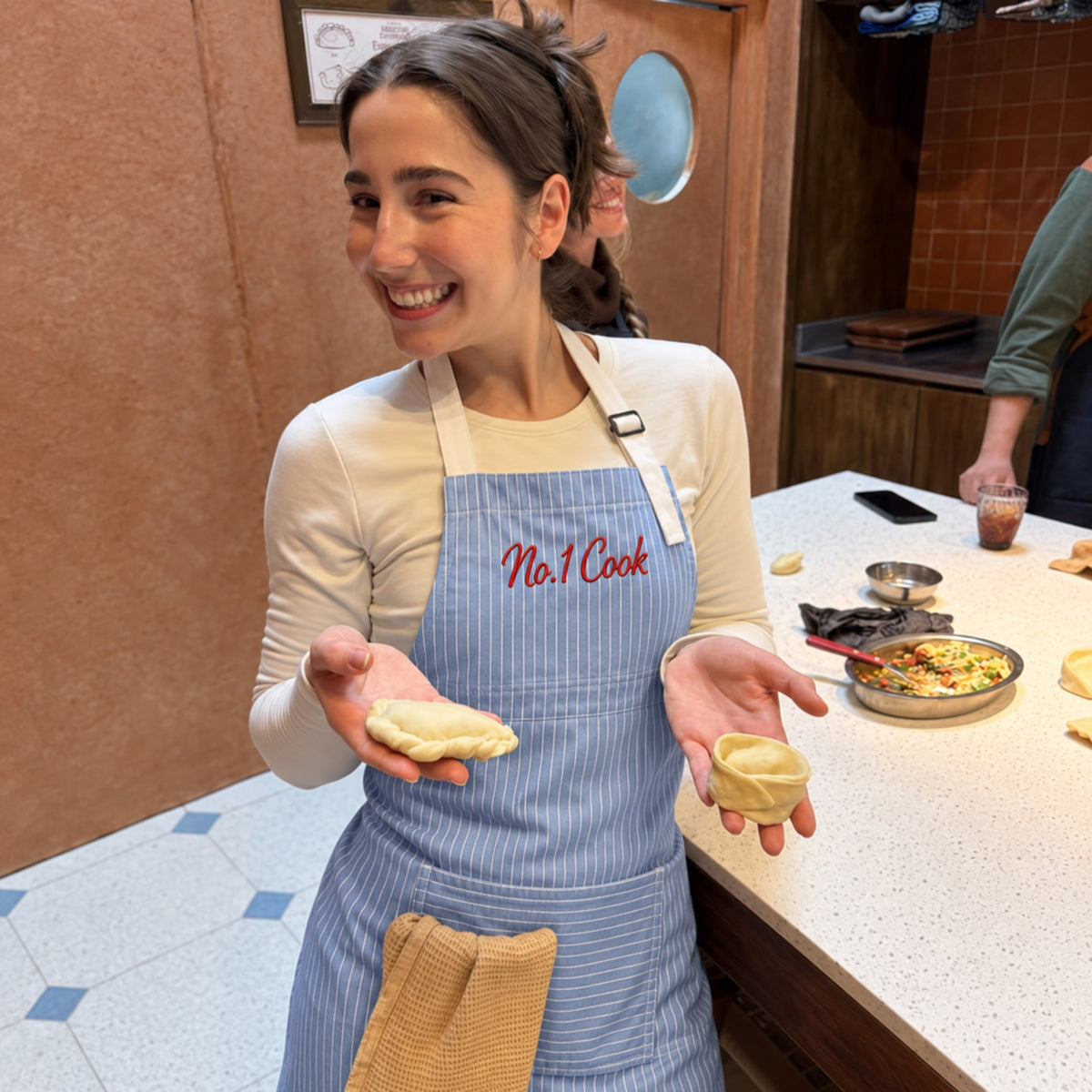 A smiling woman wearing a light blue and white striped apron with 'No.1 Cook' embroidered in red on the chest, holding two small pieces of dough in a kitchen setting.