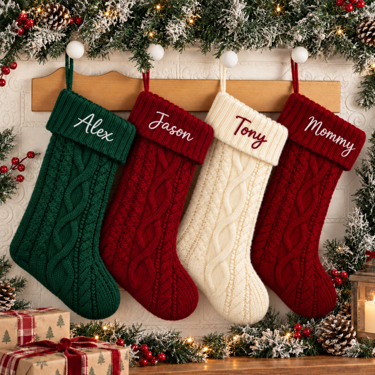 Four personalized Christmas stockings hang on a wooden rack against a white brick wall, decorated with holiday greenery and lights. The stockings are green, dark red, cream, and dark red, each with a cable-knit body and a ribbed cuff embroidered with a personalized name: Alex, Jason, Tony, and Mommy.
