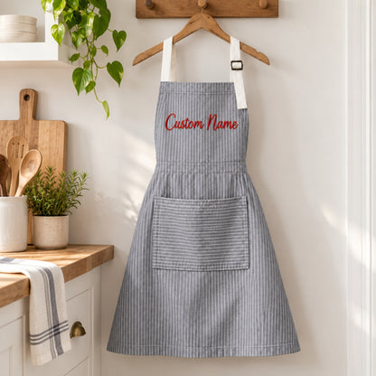 A gray and white vertically striped kitchen apron with white shoulder straps and red embroidered text reading 'Custom Name' on the chest, hanging on a wooden hanger against a white wall in a bright kitchen setting.